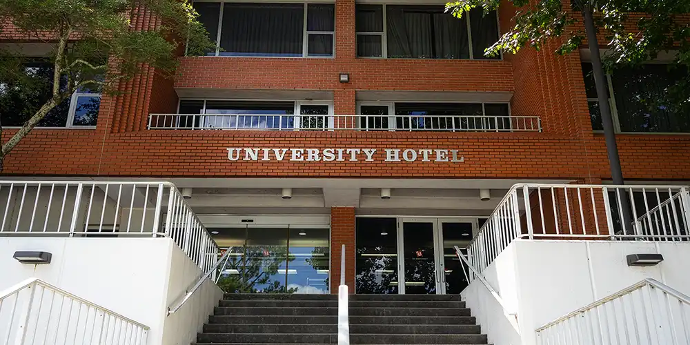 Entrance to the University Hotel, a red-brick building with the words “UNIVERSITY HOTEL” displayed above the main entrance. A central staircase leads up to glass doors, with white railings on both sides and trees framing the building facade.